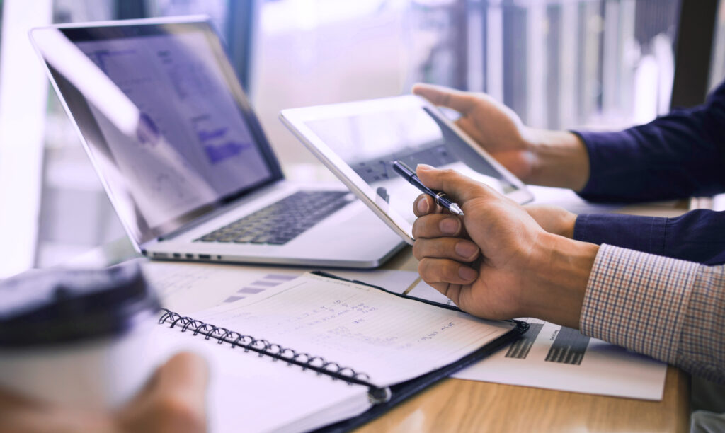 Dwie osoby pracują przy biurku z laptopem, tabletem, dokumentami i otwartym notatnikiem. Jedna trzyma długopis, druga kubek z kawą. Wykresy i dane są widoczne na ekranach i papierach, sugerując spotkanie biznesowe lub analizę.