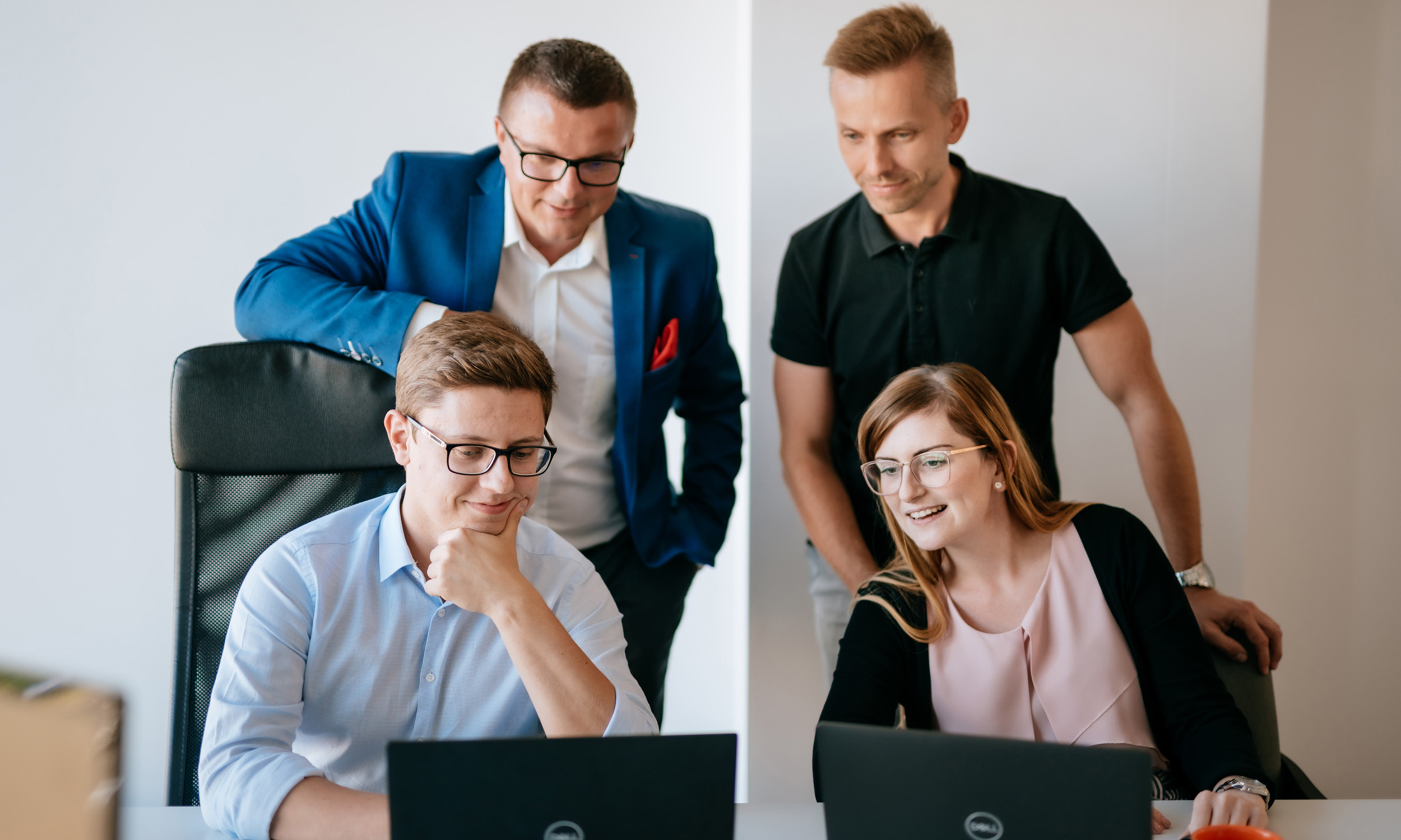 Four colleagues work together at a desk, looking at two laptops and smiling. Two people are seated while two stand behind them, all appearing engaged in a collaborative office environment.
