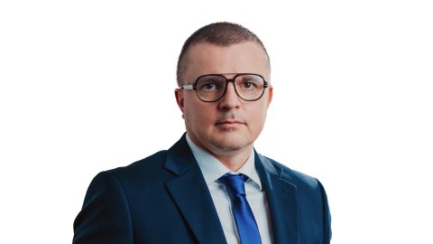 Mężczyzna w ciemnoniebieskim garniturze, białej koszuli, niebieskim krawacie i dużych okularach, z krótkimi włosami, stoi na prostym białym tle, uosabiając profesjonalizm niezbędny w cyfrowej transformacji sprzedaży B2B.