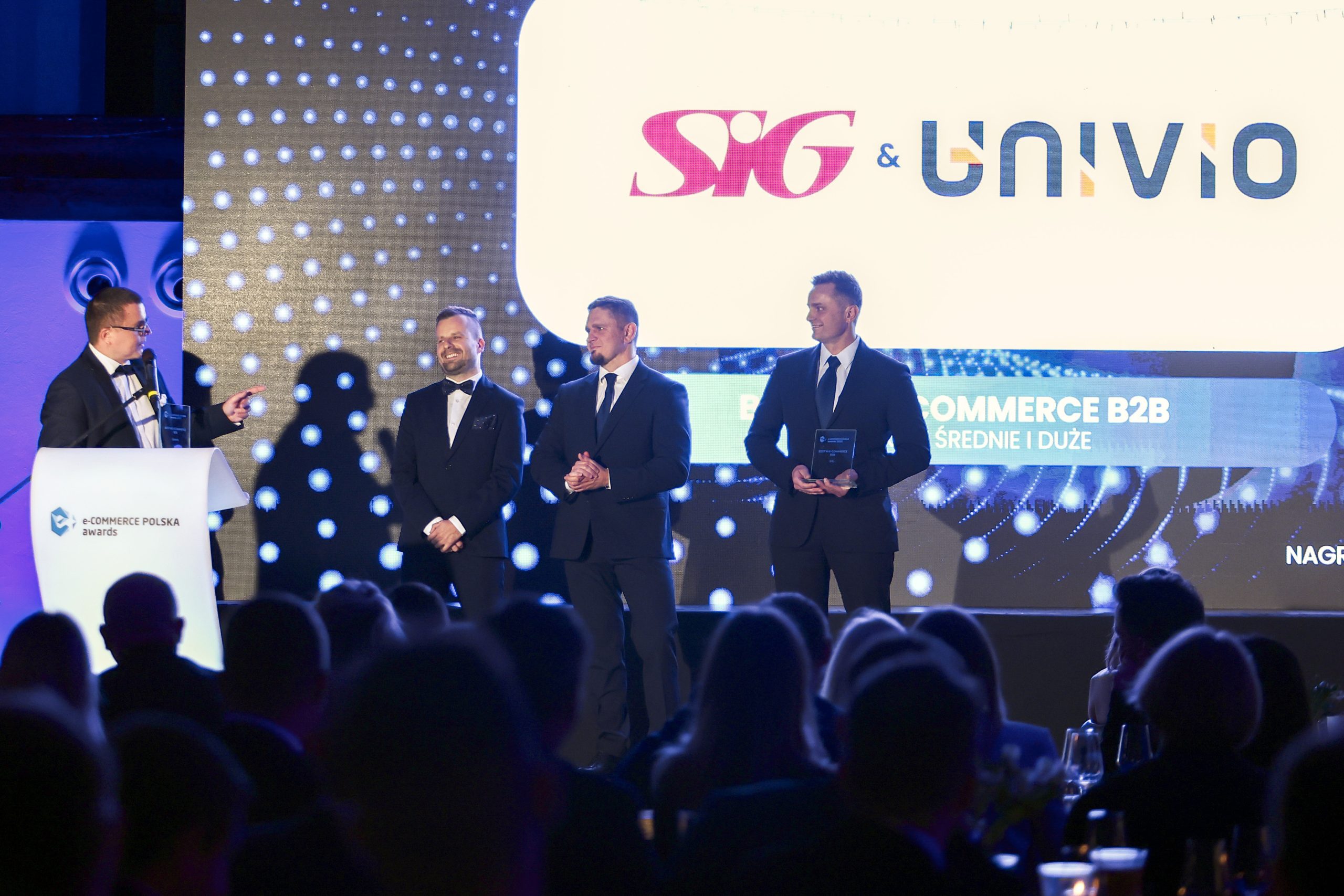 Czterech mężczyzn w garniturach stoi na scenie podczas rozdania nagród e-commerce B2B w branży budowlanej. Jeden z nich mówi do mikrofonu, podczas gdy pozostali słuchają. Duży ekran za nimi wyświetla logo SIG i UNIVIO oraz szczegóły wydarzenia.