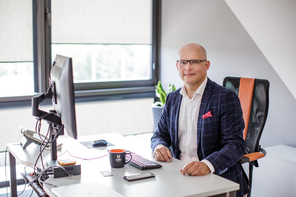 Mężczyzna w biznesowym garniturze siedzi przy biurku w nowoczesnym biurze, uśmiechając się do kamery. Korzysta z komputera, a na biurku leży telefon, kubek z kawą i notatnik. Duże okna wpuszczają naturalne światło.