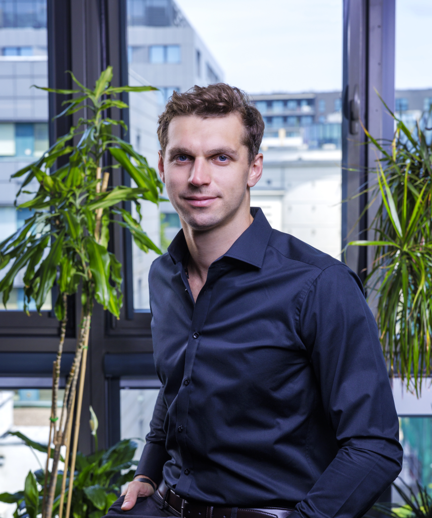 A man in a dark blue button-up shirt stands indoors near tall green plants, with large windows and modern buildings visible in the background.