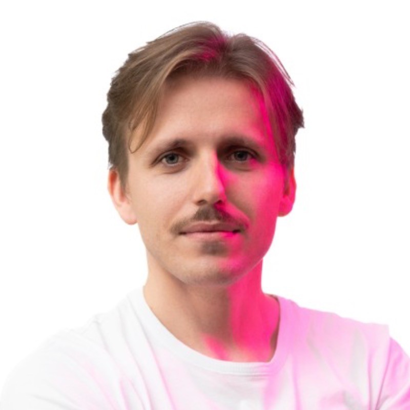 Mężczyzna z jasnobrązowymi włosami i wąsami, ubrany w białą koszulę, stoi na białym tle z różowym światłem rzucającym poświatę na prawą stronę jego twarzy.