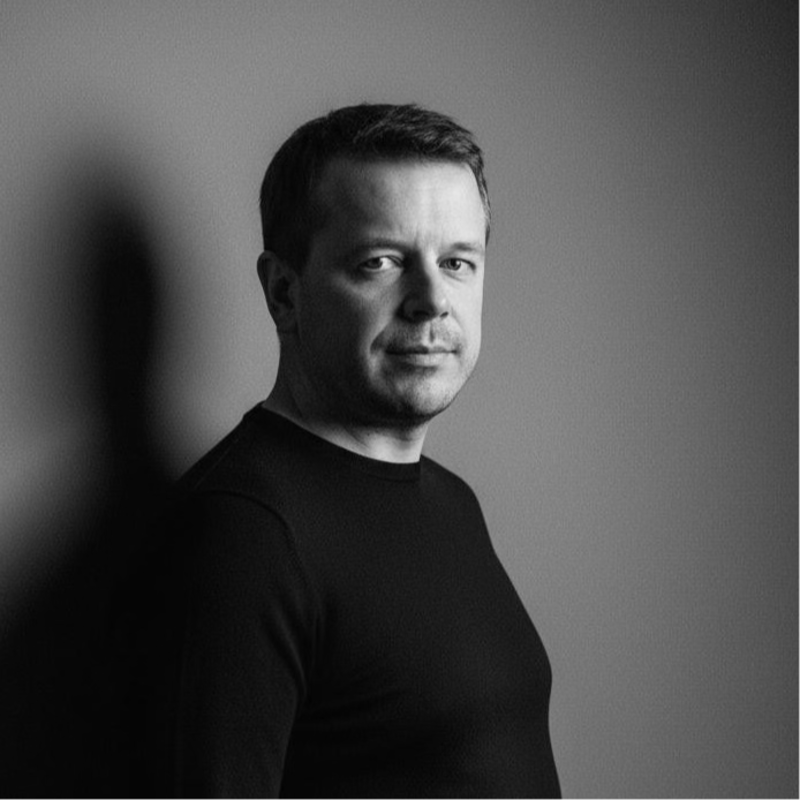 Mężczyzna z krótkimi włosami, ubrany w ciemną koszulę z długim rękawem, stoi na prostym tle z miękkim oświetleniem, rzucającym cień na ścianę za nim. Patrzy spokojnie w stronę kamery.