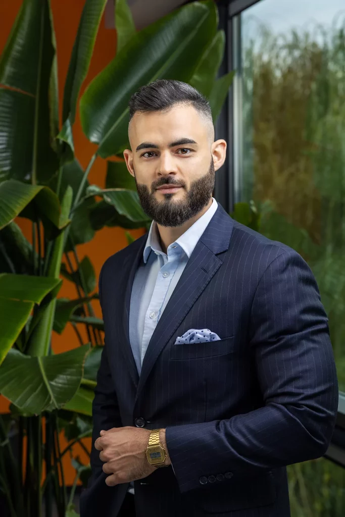 Mężczyzna z krótką brodą i starannie ułożonymi włosami ma na sobie ciemny garnitur w prążki, jasnoniebieską koszulę i złoty zegarek, stojąc pewnie przed dużymi zielonymi roślinami w pobliżu okna.