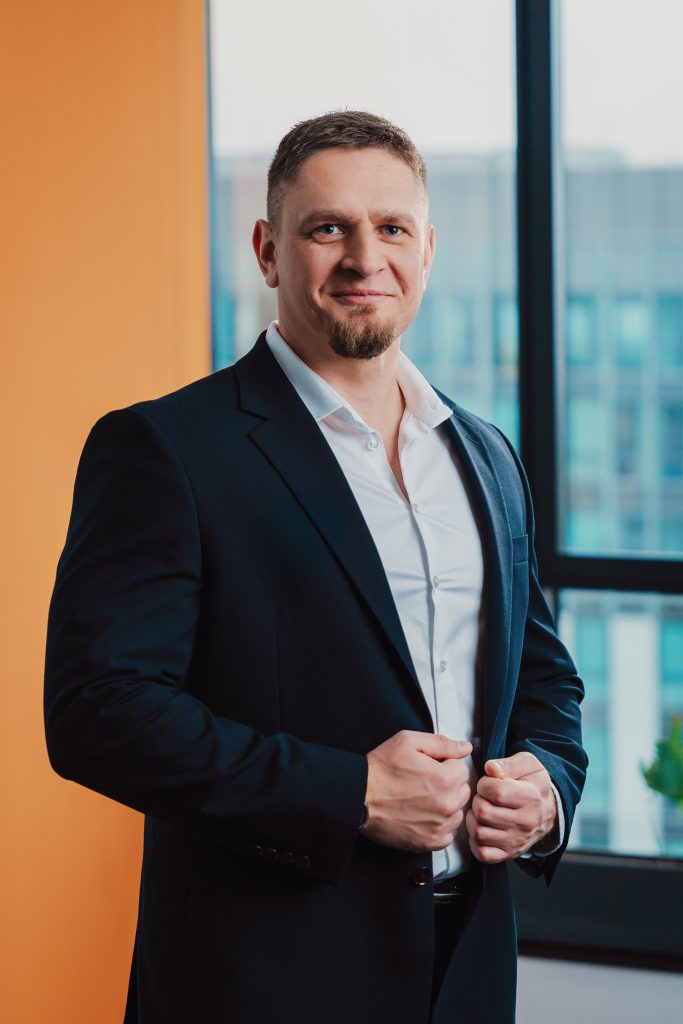 Mężczyzna ubrany w ciemny garnitur i białą koszulę stoi w pomieszczeniu w pobliżu okna, uśmiechając się pewnie z rękami trzymającymi marynarkę. Tło obejmuje pomarańczową ścianę i budynek biurowy widoczny przez okno.